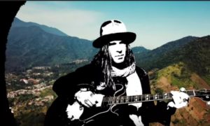 A man in a hat with a guitar in front of mountains.